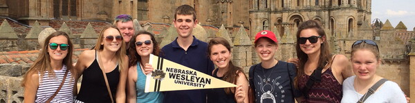 Blind student in the middle holds a Nebraska Wesleyan pennant surrounded by other students in front of a castle.