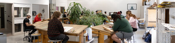 Art students drawing and sitting at large wooden drawing desks in circle around plants.