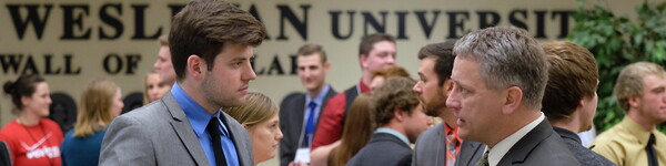 In a crowded Great Hall dressed in professional attire, a male student talks with an adult male.