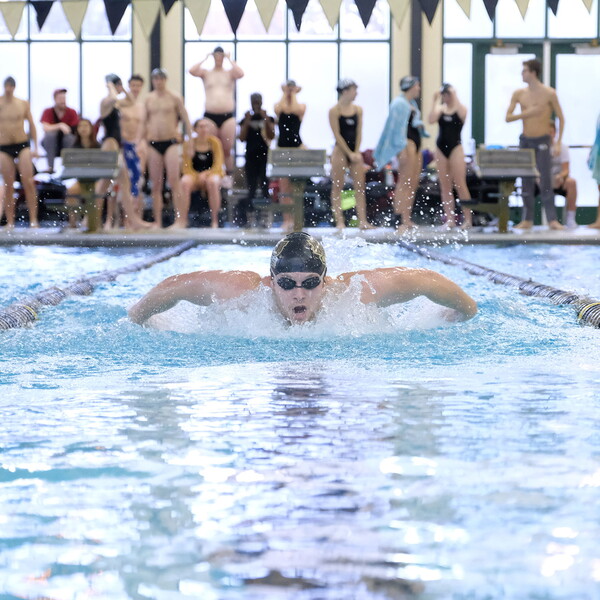 A male swimmer doing the breast stroke with a crowd of onlookers cheering him on the side of the pool behind him.