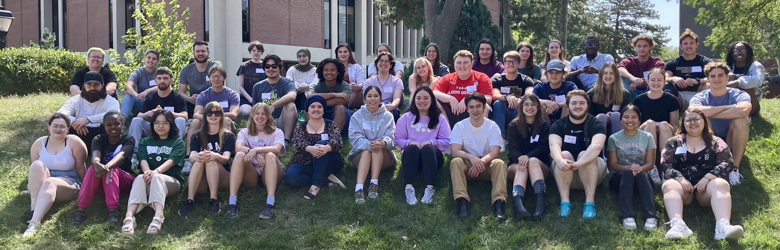 About three dozen students sit on the lawn in three rows smiling.