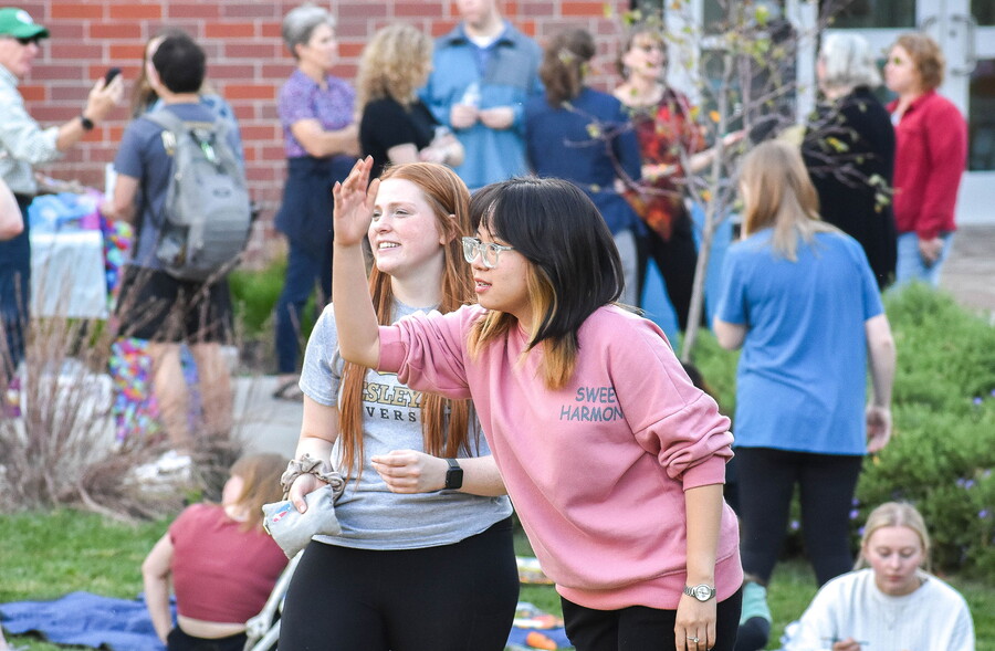 Two young women tossing a sand bag amongst many people outdoors.