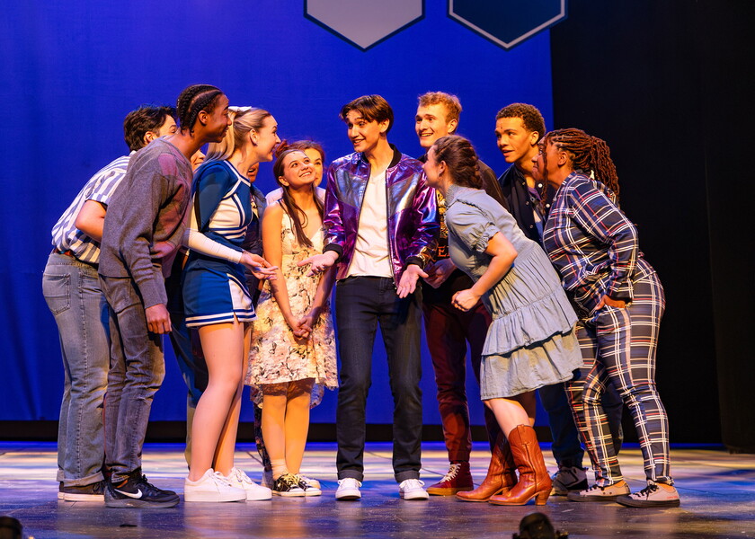 Actors on stage surround one male and bend closer to him as he delivers his lines.