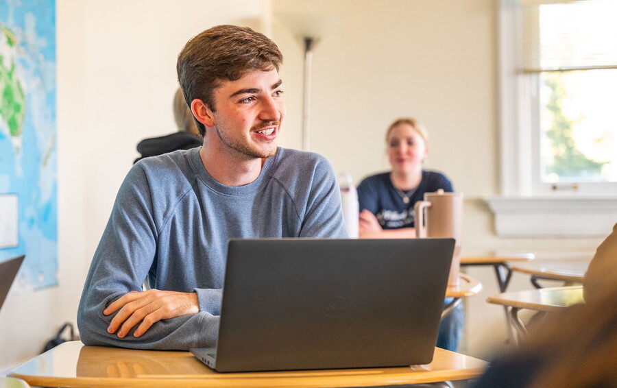 Male student sitting at a desk in a classroom with his laptop open.