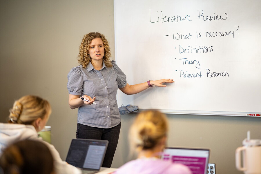 Chelsea Klinkebiel teaching in front of a class pointing towards a white board.