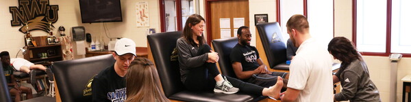In the athletic training room, Professor Wilson sits while a student wraps her ankle.