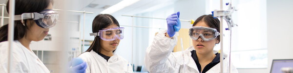 Three female students in lab coats, gloves and eyewear conduct an experiment in chemistry class.