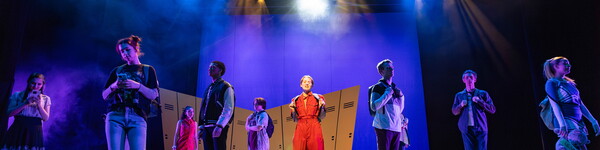 Several actors on the stage standing in front of set with lockers singing.