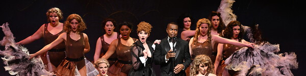 A man in a tux, a woman in a gown and dancers with large feather fans perform on stage.